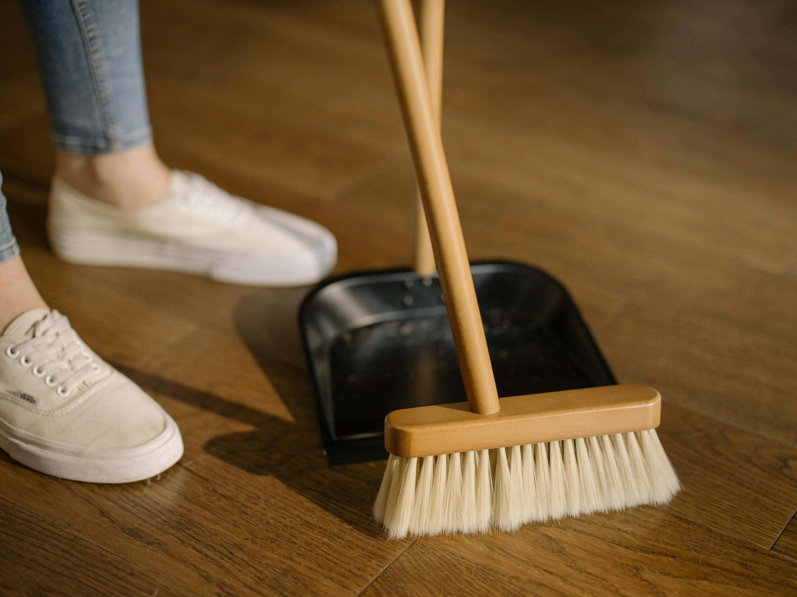 Close-up of cleaning process with broom and dustpan beside sneakers on a wooden indoor floor.